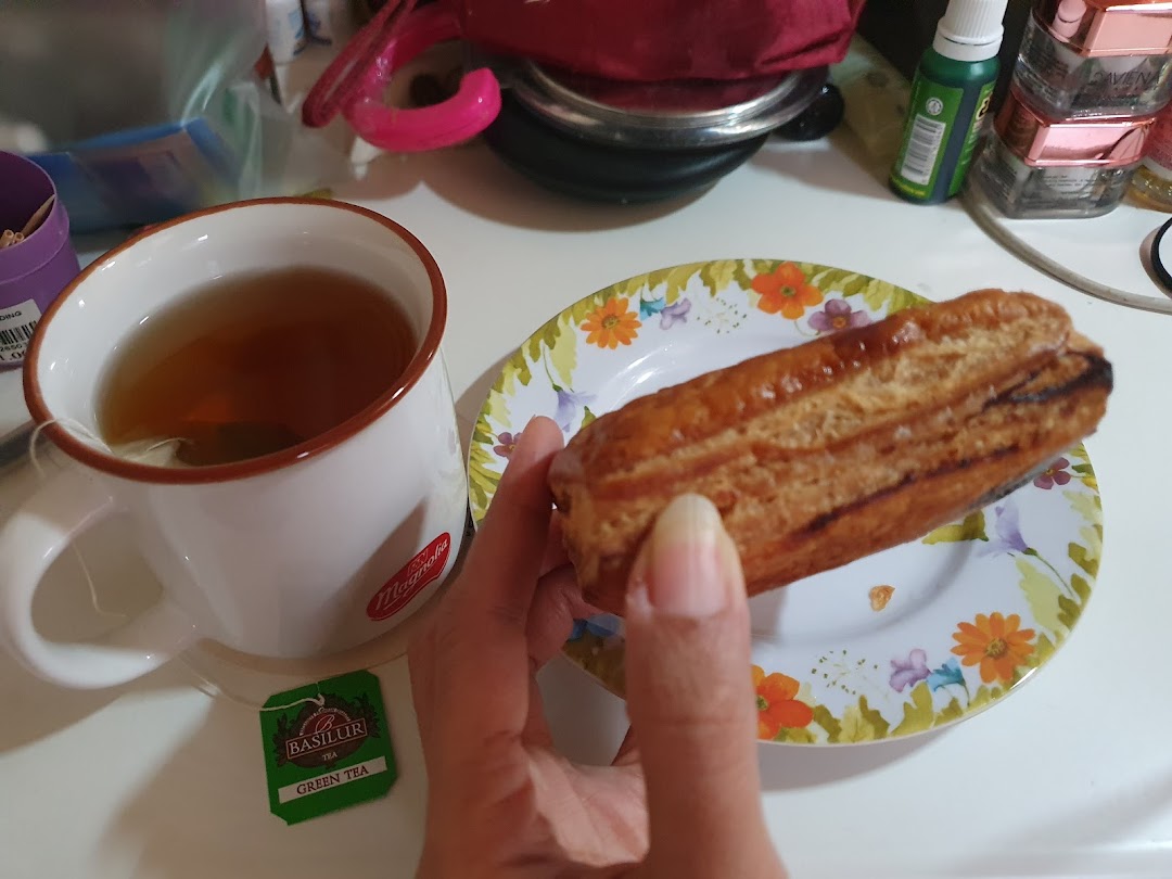 A hand holds a golden pastry next to a cup of tea and a tea bag on a floral plate.