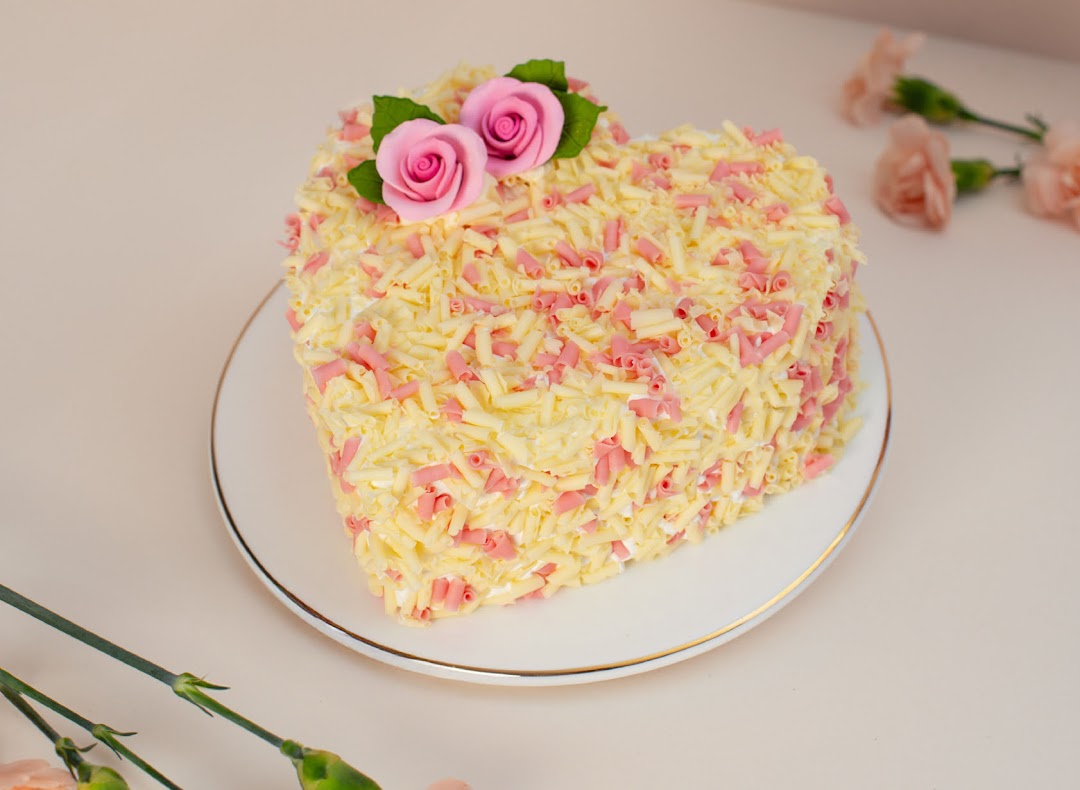 A heart-shaped cake adorned with white and pink chocolate shavings and delicate pink rose decorations.