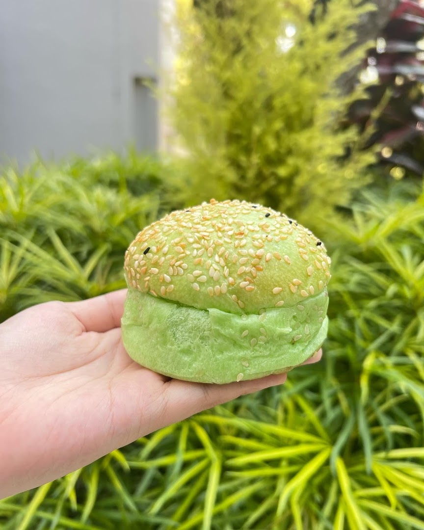 A hand holds a vibrant green bun topped with sesame seeds against a lush green foliage background.