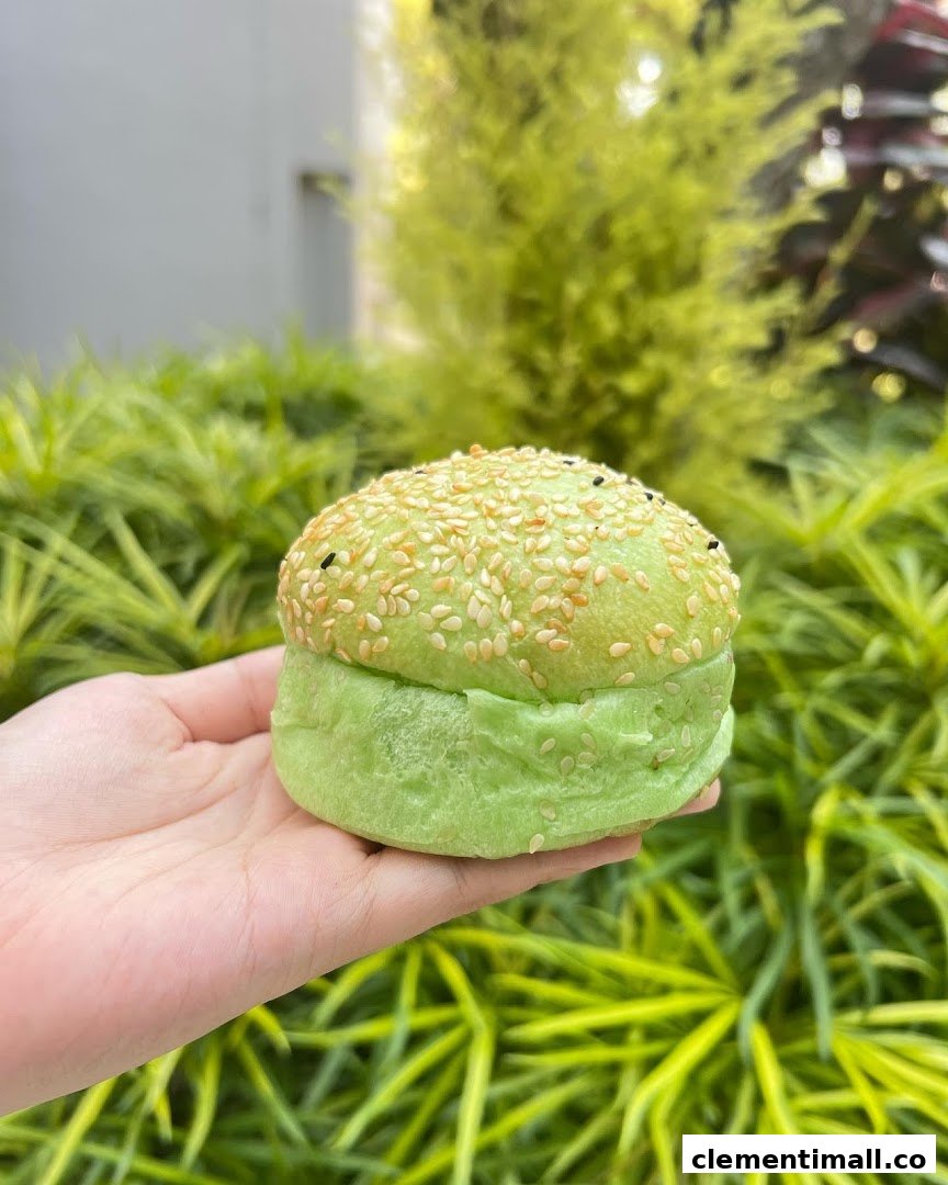 A hand holds a vibrant green bun topped with sesame seeds against a lush green foliage background.