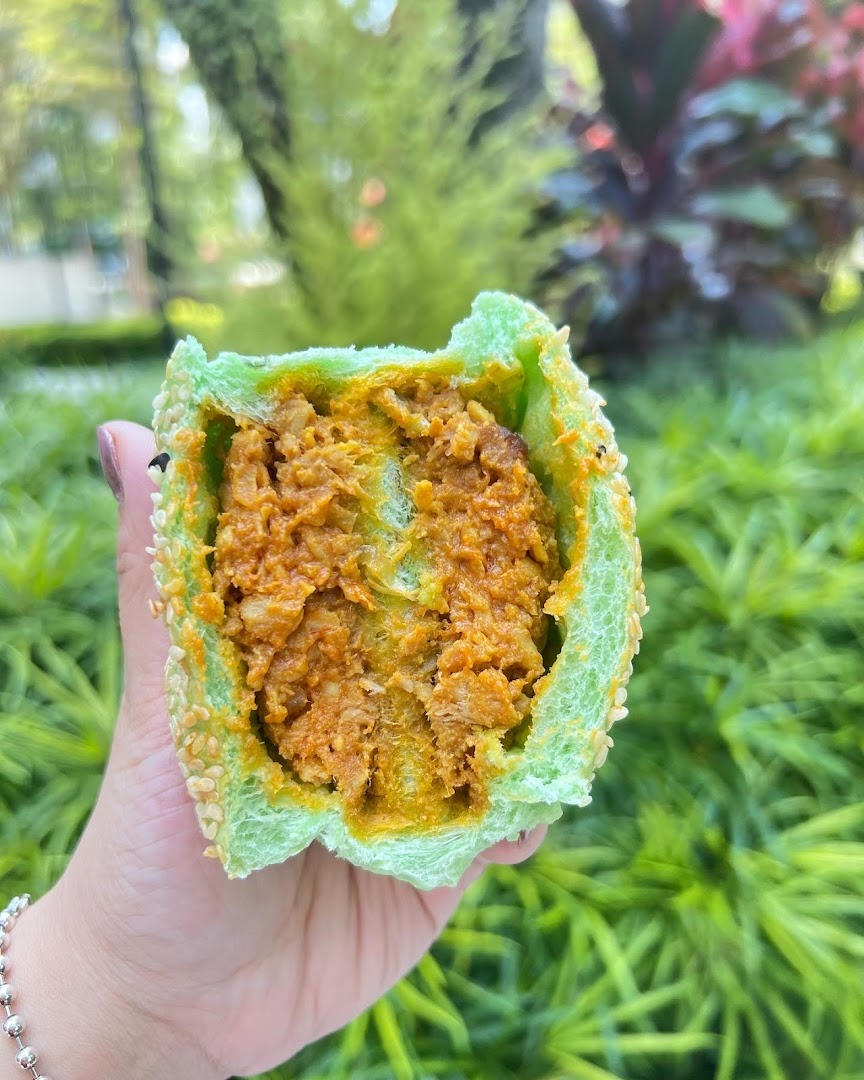 A hand holds a vibrant green bun filled with savory orange curry, set against a lush green background.