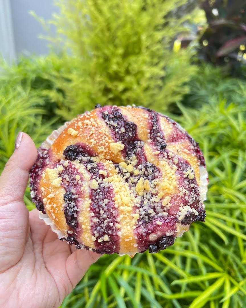 A hand holds a freshly baked blueberry crumble bun against a vibrant green foliage background.