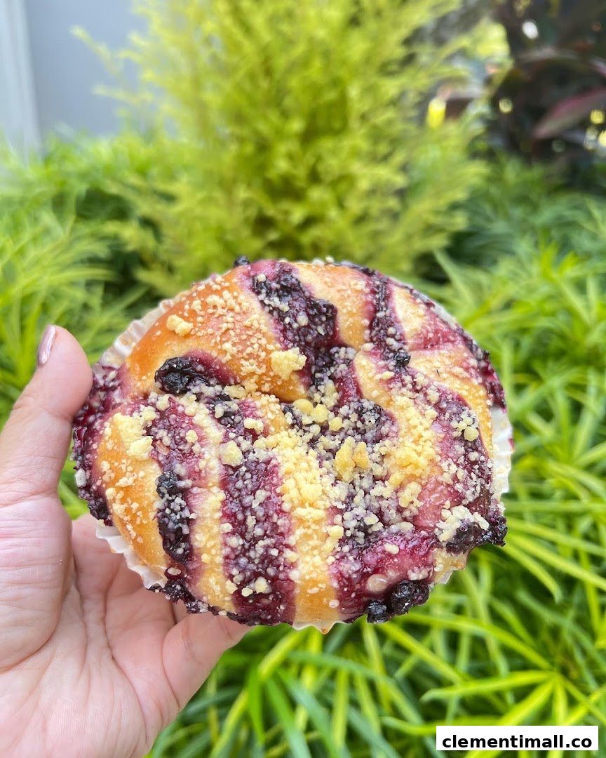 A hand holds a freshly baked blueberry crumble bun against a vibrant green foliage background.