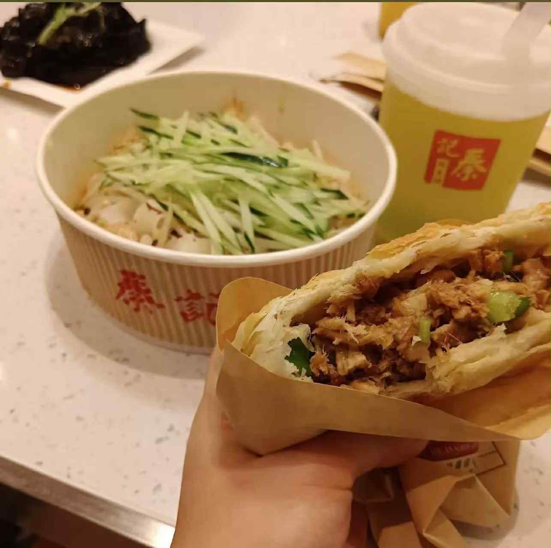 A hand holds a savory rougamo, with a bowl of noodles and a refreshing drink in the background.