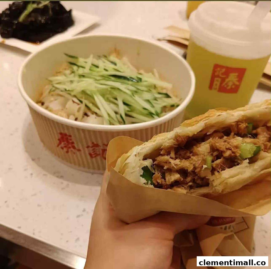 A hand holds a savory rougamo, with a bowl of noodles and a refreshing drink in the background.