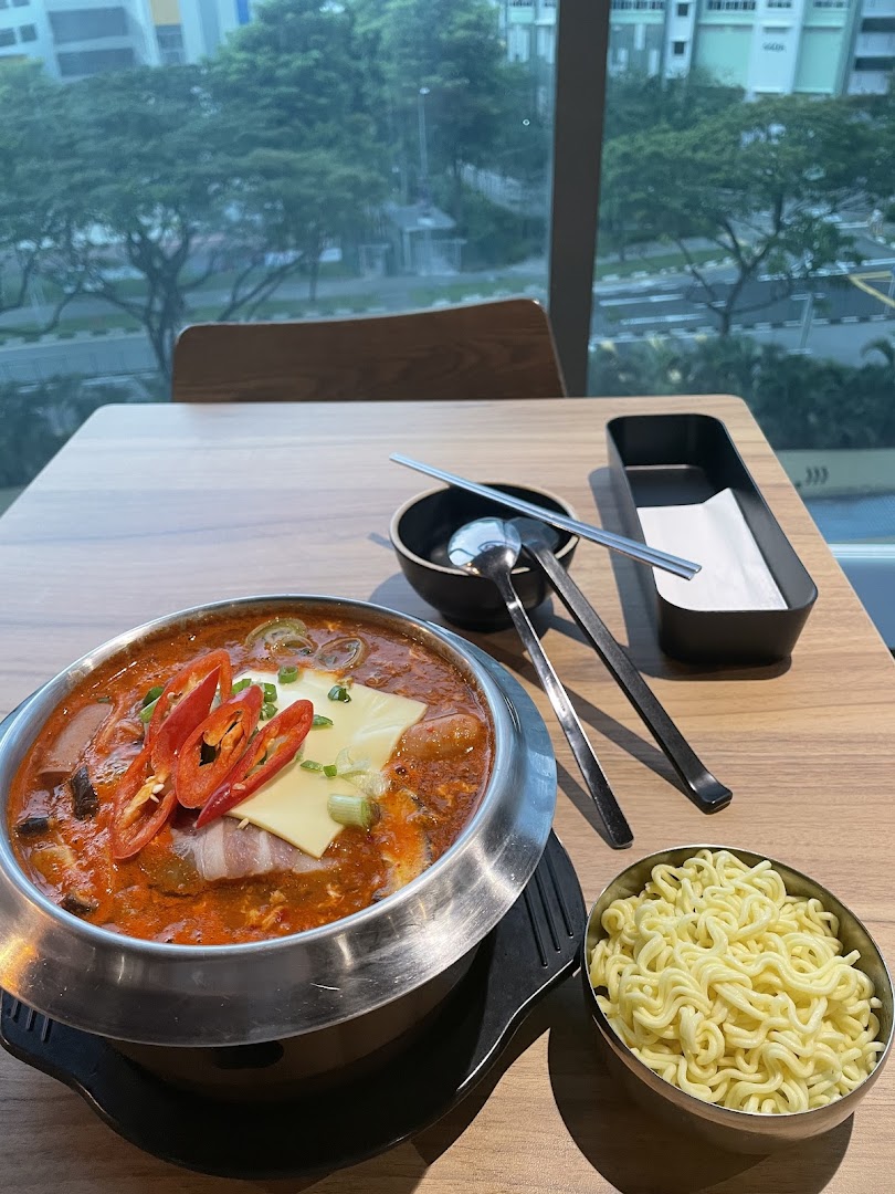 A vibrant Korean hot pot dish with noodles and cheese, served by a window with a view.