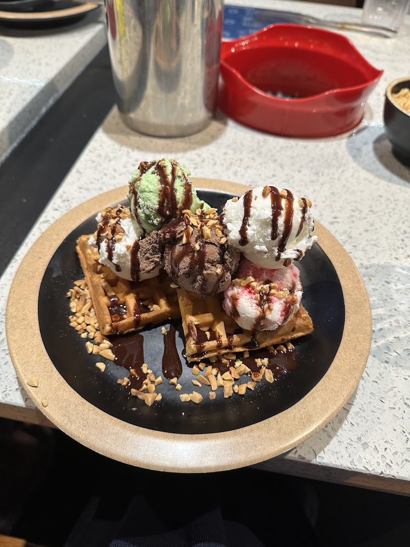 Waffles topped with multiple scoops of ice cream, chocolate sauce, and nuts on a black plate.