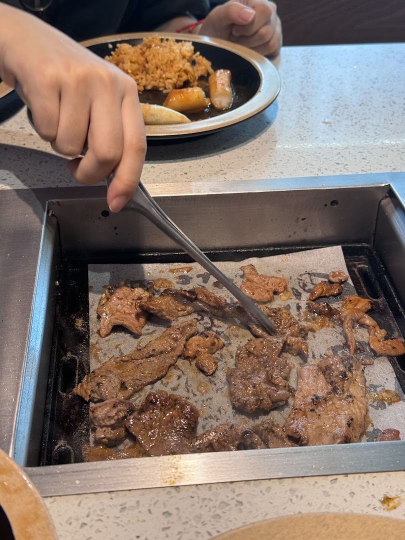 A person grilling marinated meat on a tabletop BBQ grill, with a plate of fried rice and side dishes nearby.
