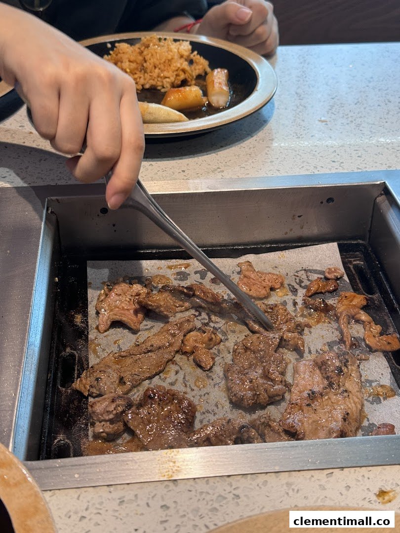 A person grilling marinated meat on a tabletop BBQ grill, with a plate of fried rice and side dishes nearby.