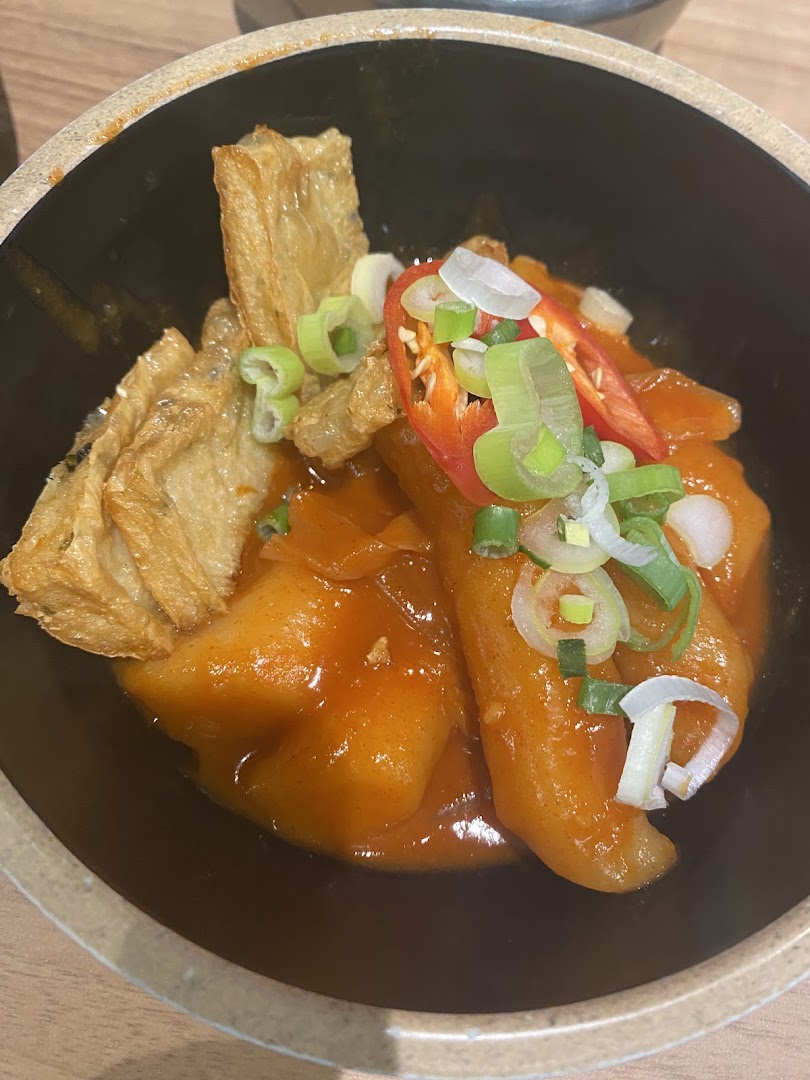 A close-up of a vibrant Korean dish with rice cakes, beancurd skin, chili, and spring onions in a rich sauce.