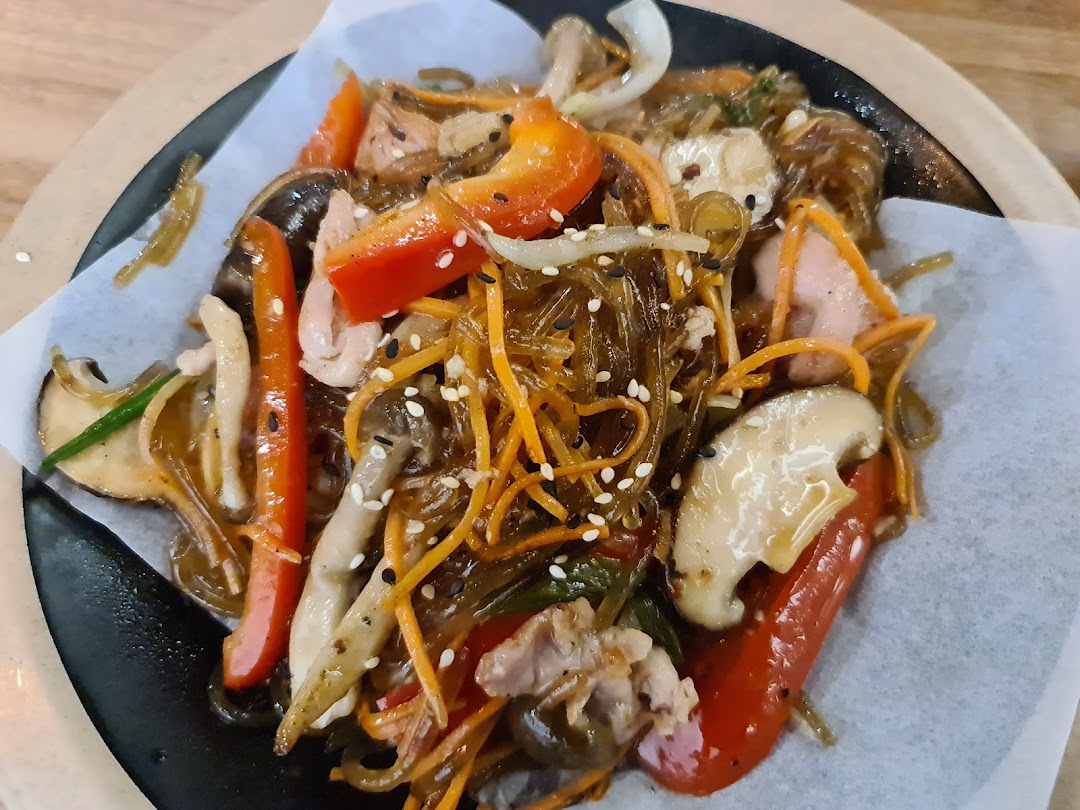A vibrant close-up of a Korean glass noodle dish (Japchae) with vegetables, mushrooms, and meat.