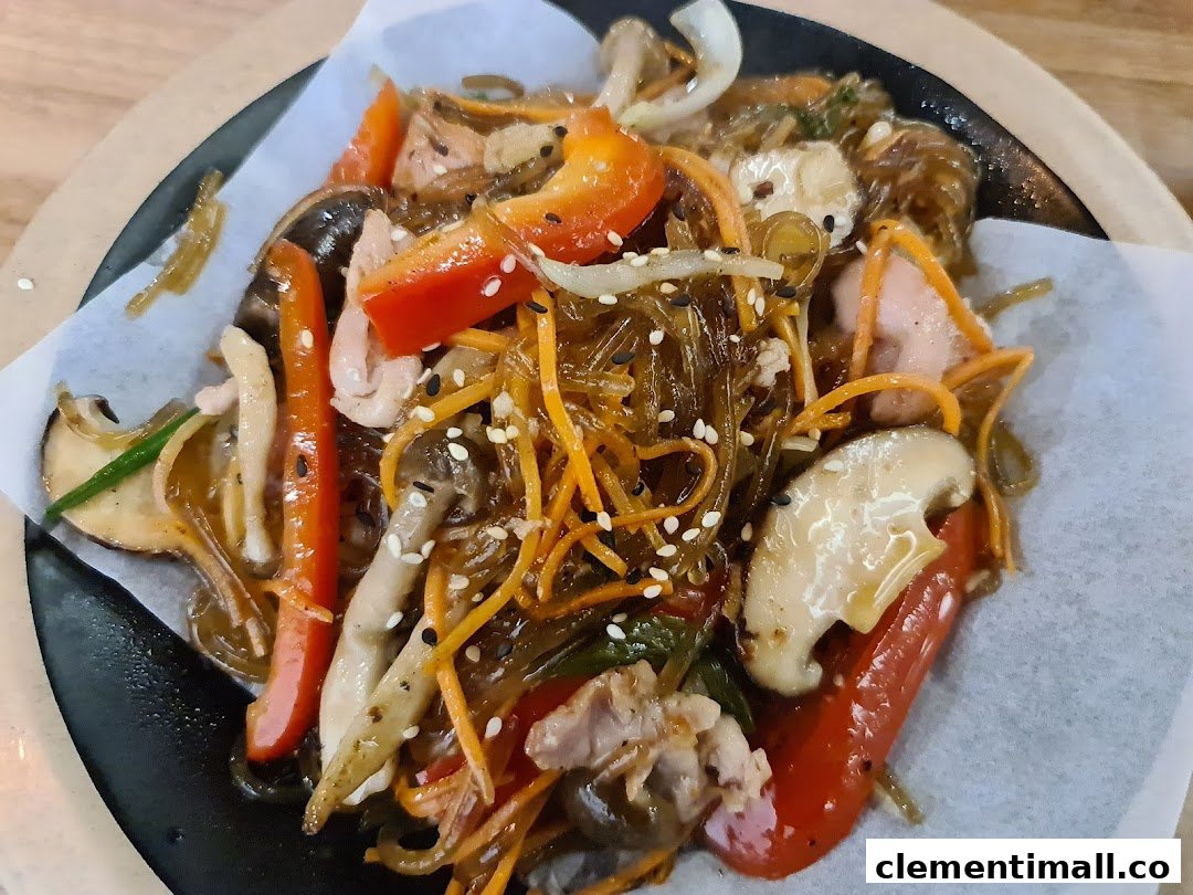 A vibrant close-up of a Korean glass noodle dish (Japchae) with vegetables, mushrooms, and meat.