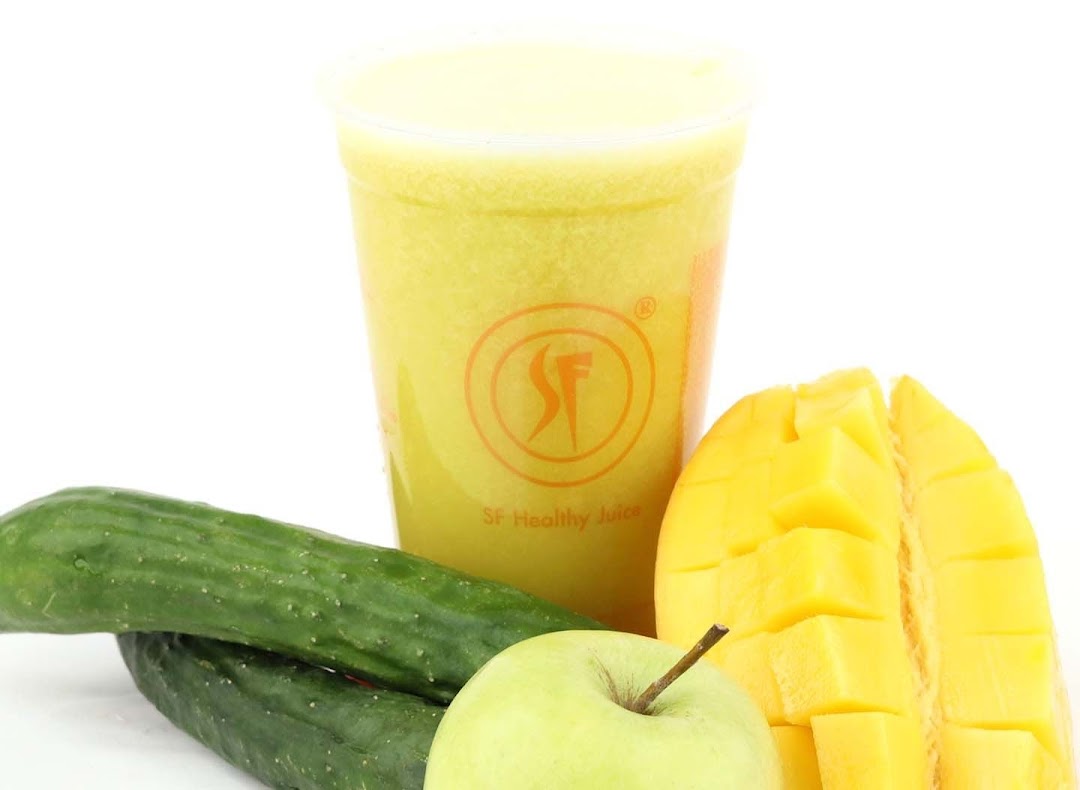 A vibrant yellow juice in a branded cup, surrounded by fresh mango, cucumber, and green apple.