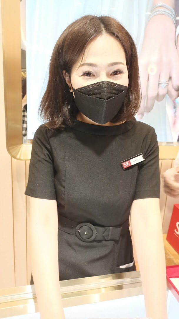 A friendly staff member from SK Jewellery, wearing a black uniform and mask, smiles behind a counter.