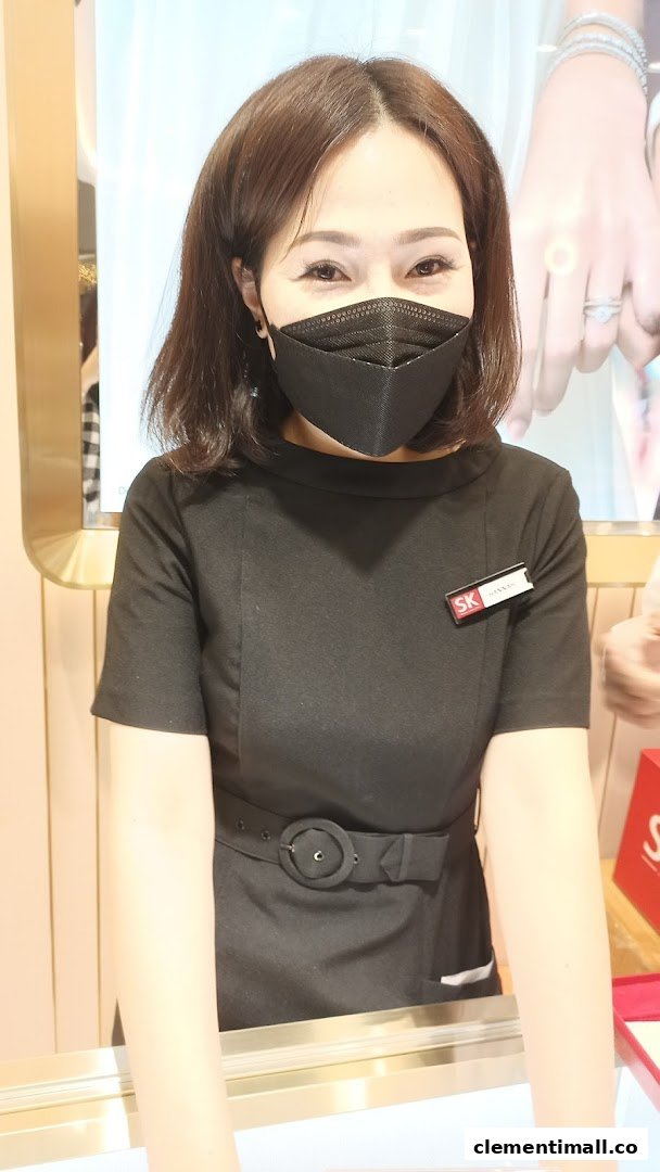 A friendly staff member from SK Jewellery, wearing a black uniform and mask, smiles behind a counter.