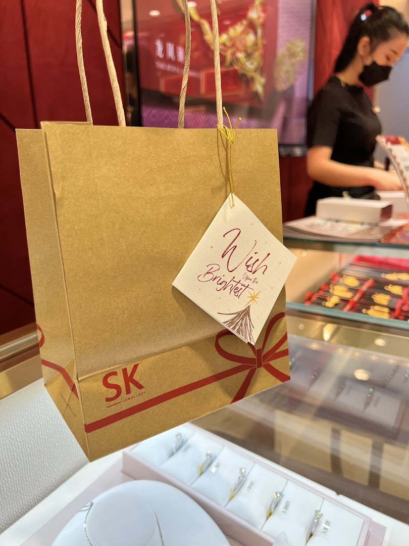A branded shopping bag with a gift tag in an SK Jewellery store, featuring displays and a staff member.