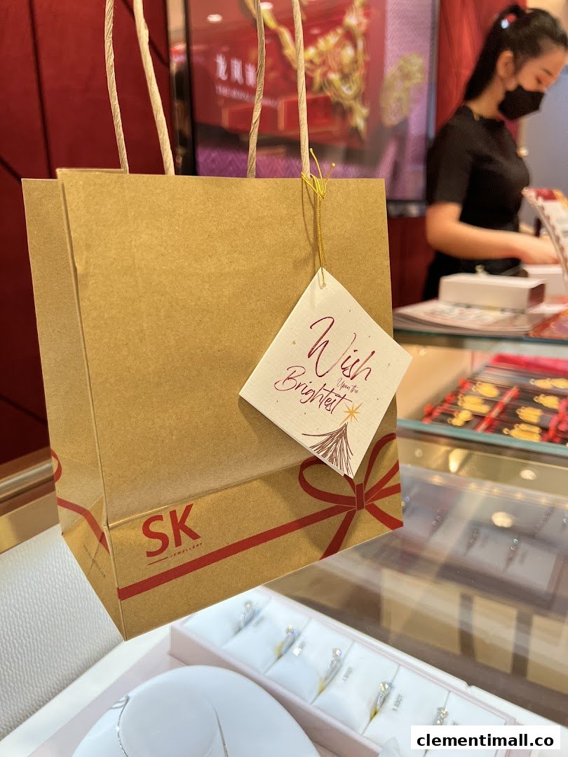 A branded shopping bag with a gift tag in an SK Jewellery store, featuring displays and a staff member.