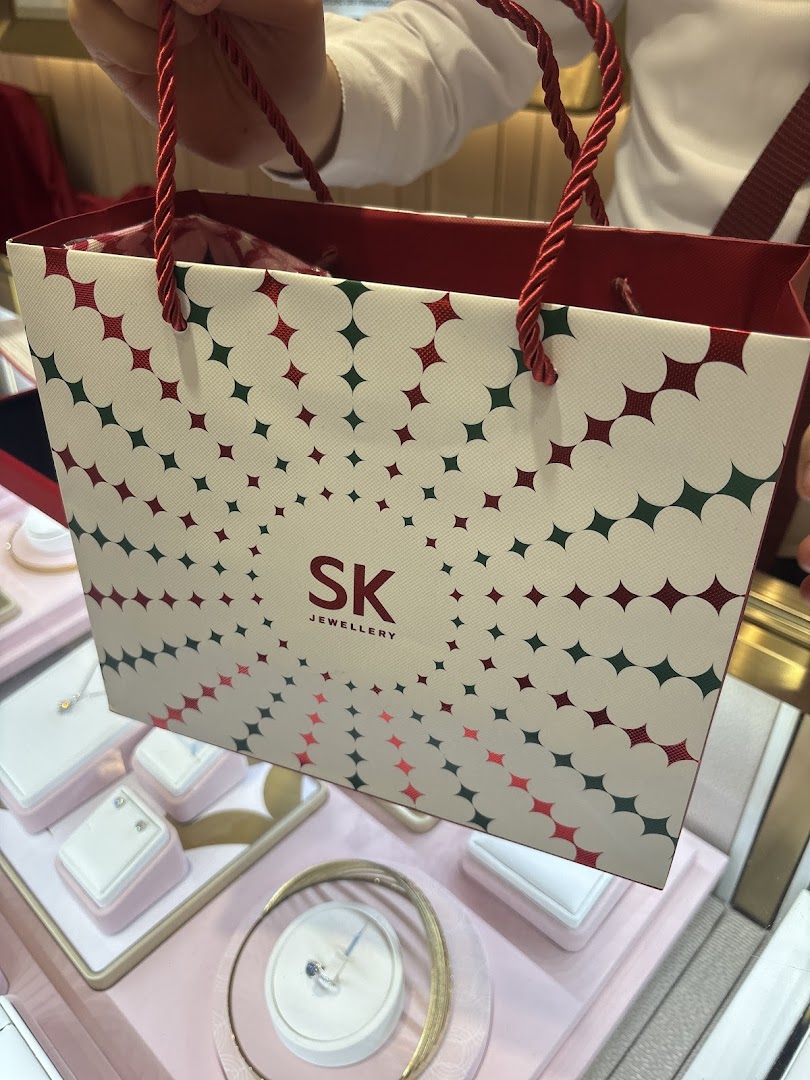 A person holds an SK Jewellery shopping bag, with elegant jewellery displays visible in the background.