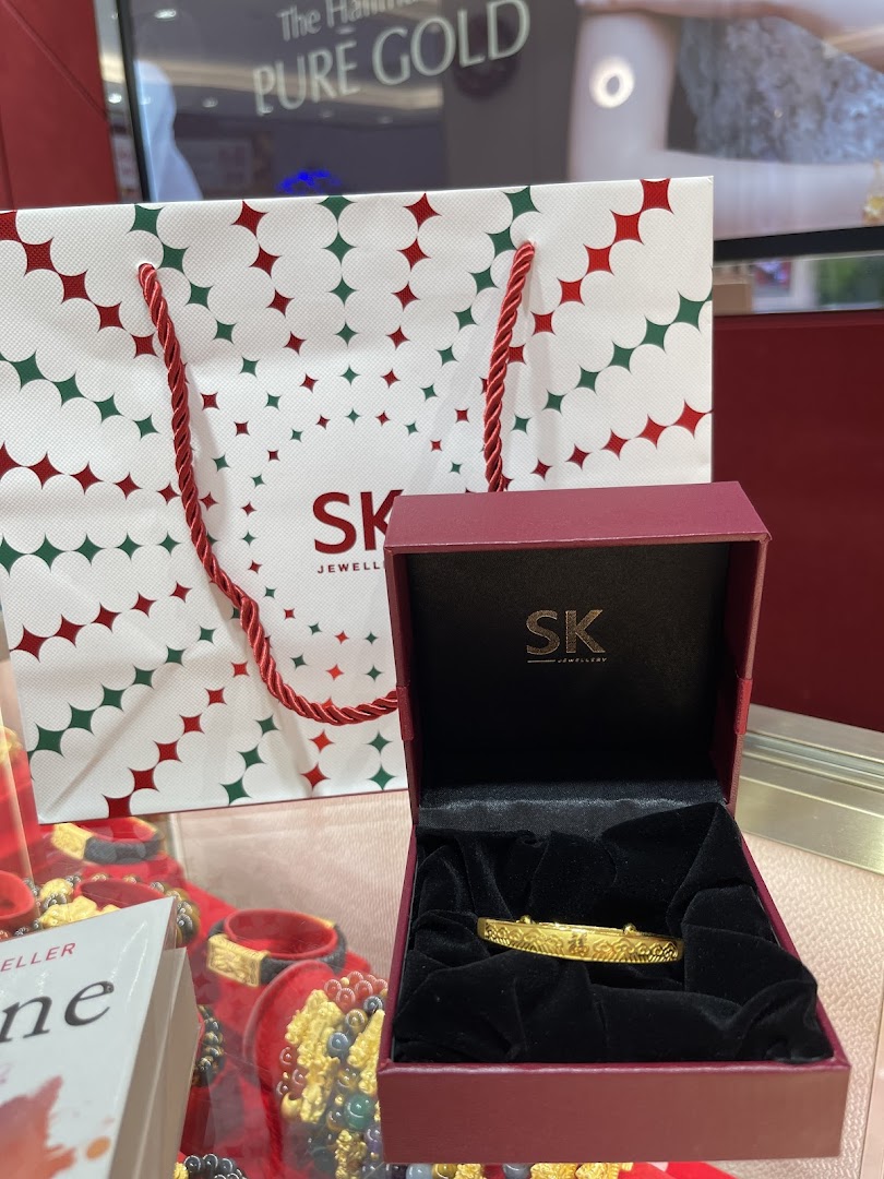 A gold bracelet in an open SK Jewellery box with a branded shopping bag and display.