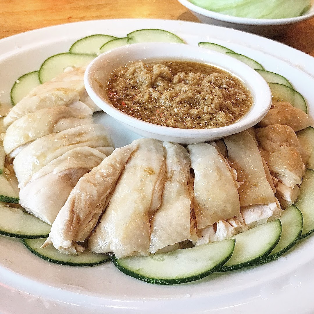 A plate of Hainanese steamed chicken with cucumber slices and a savory ginger dipping sauce.