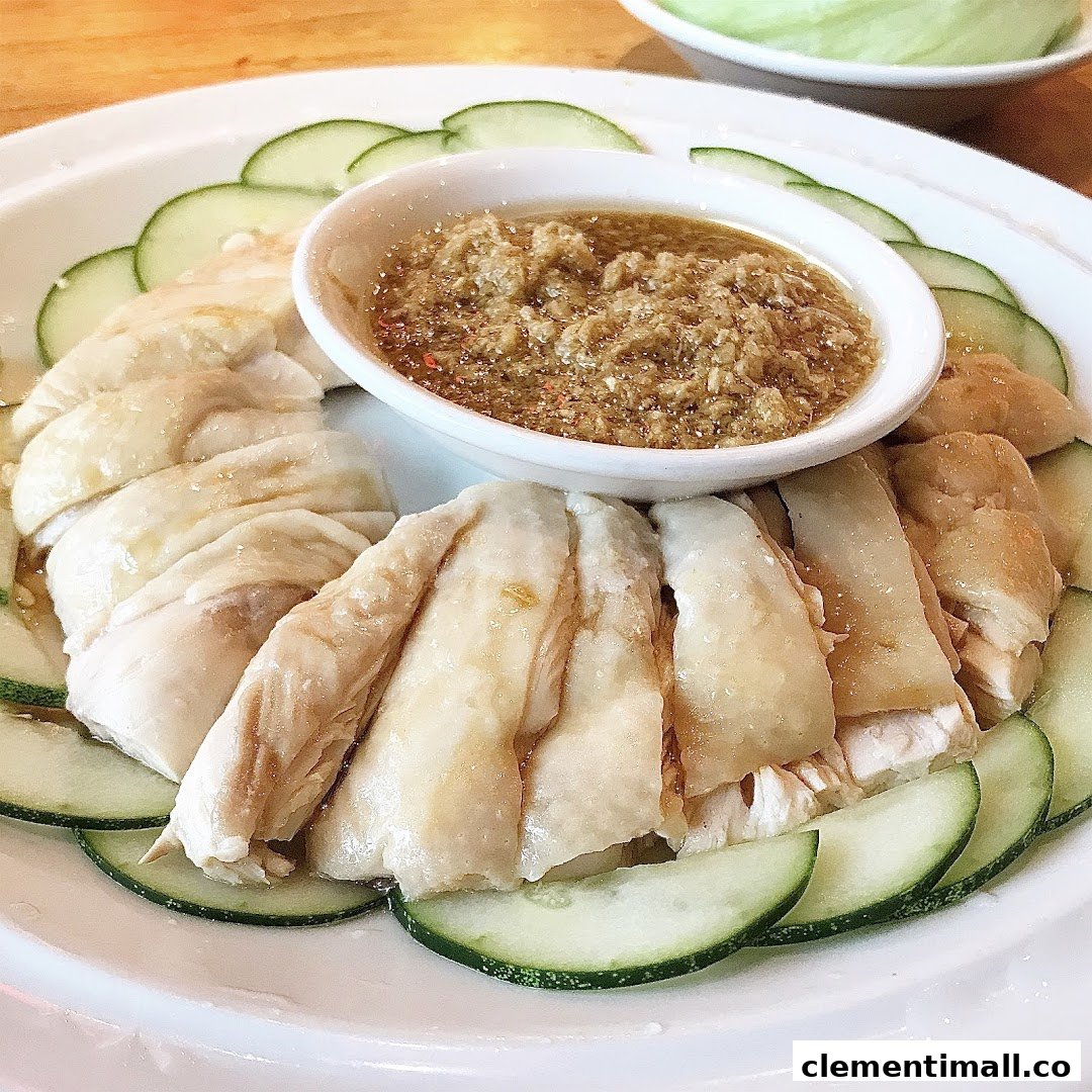 A plate of Hainanese steamed chicken with cucumber slices and a savory ginger dipping sauce.