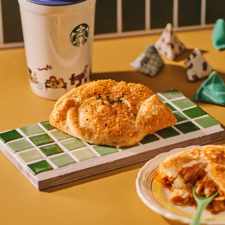 A golden-brown pastry on a green tiled board, with a Starbucks tumbler and another pastry on a plate.