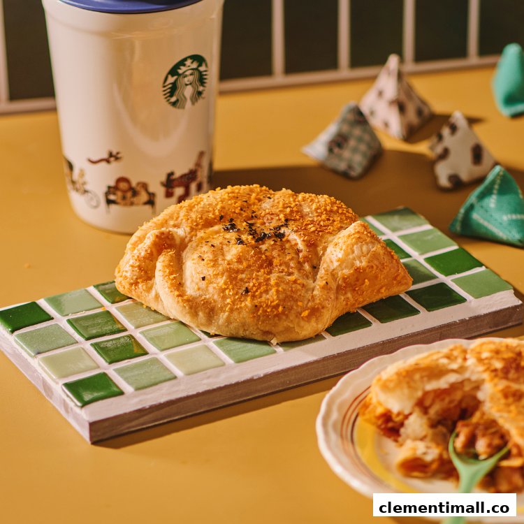 A golden-brown pastry on a green tiled board, with a Starbucks tumbler and another pastry on a plate.