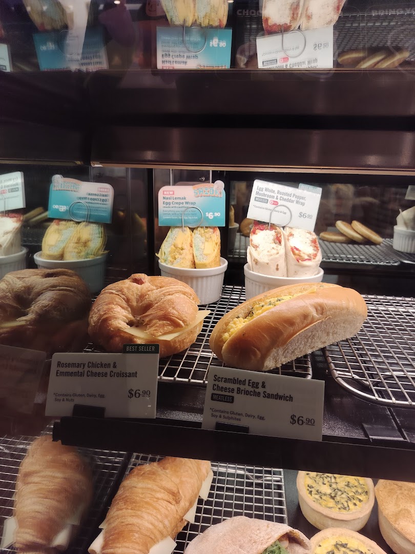 A display case at Starbucks showcasing a variety of fresh pastries, wraps, and sandwiches with prices.