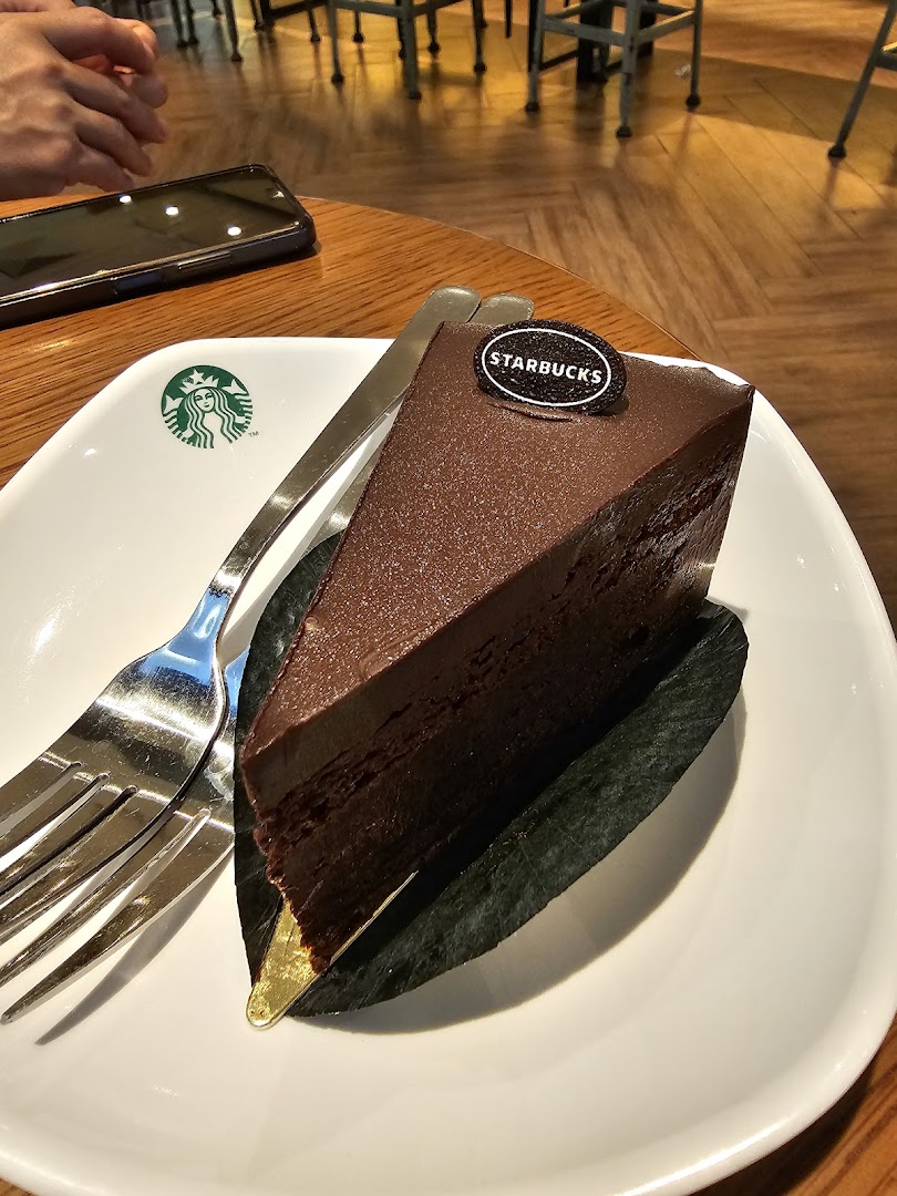 A rich chocolate cake slice on a Starbucks plate with a fork and phone on a wooden table.