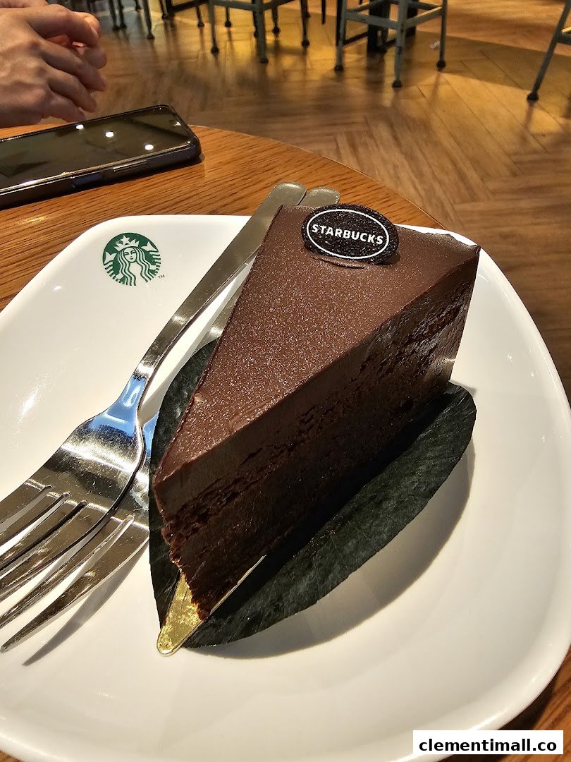 A rich chocolate cake slice on a Starbucks plate with a fork and phone on a wooden table.