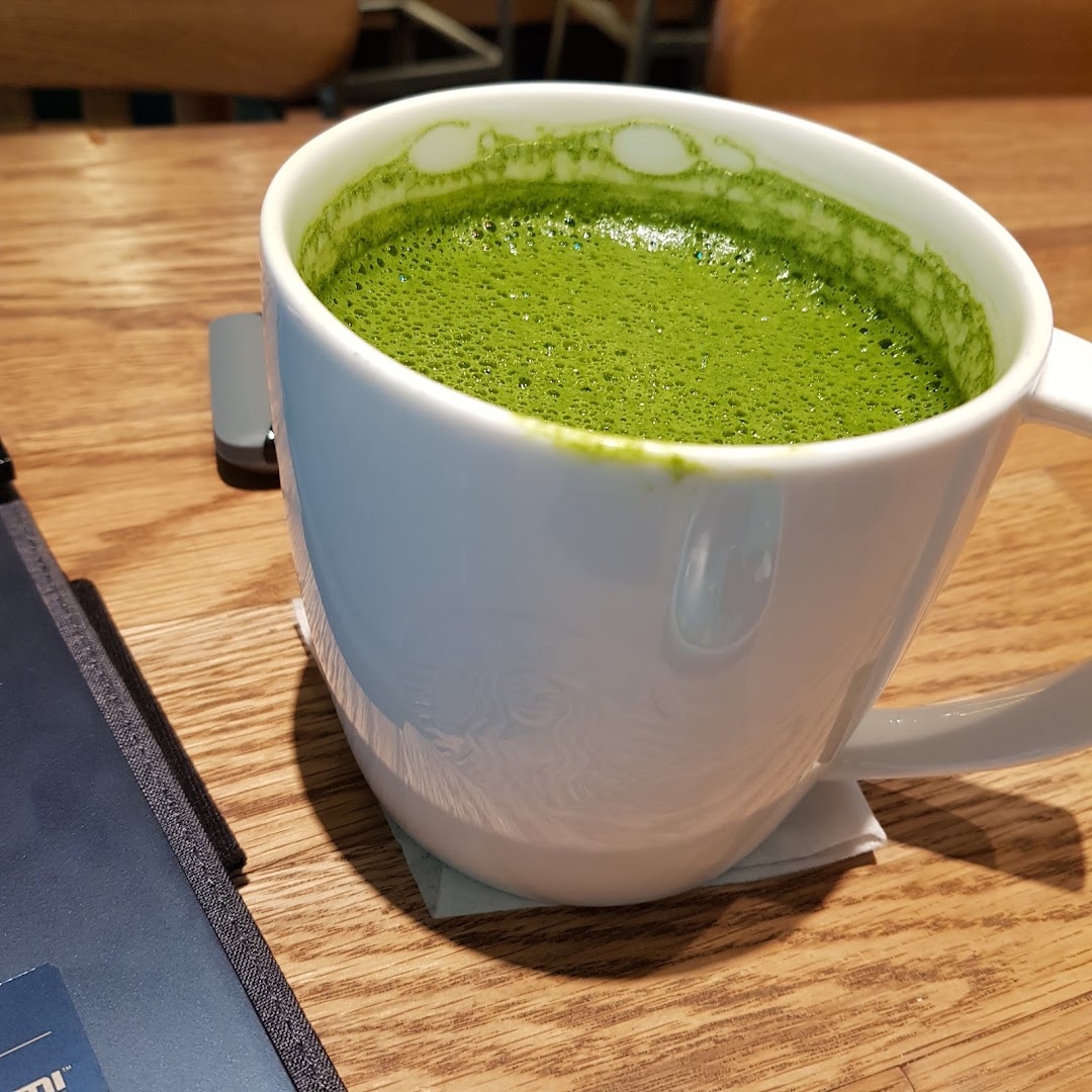 A close-up of a frothy green matcha latte in a white mug on a wooden table at Starbucks.