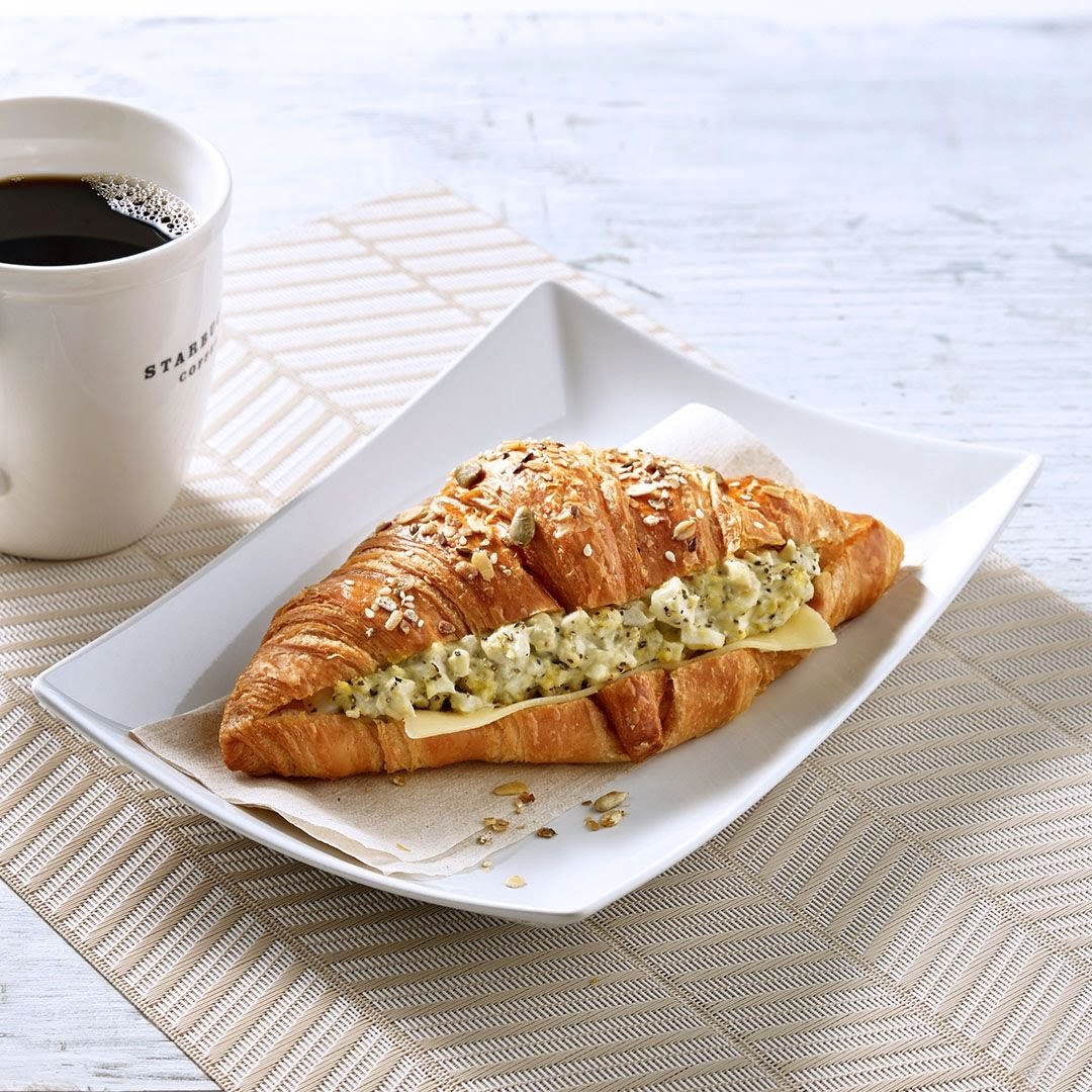 A delicious croissant sandwich filled with egg salad and cheese, served with a cup of Starbucks coffee.