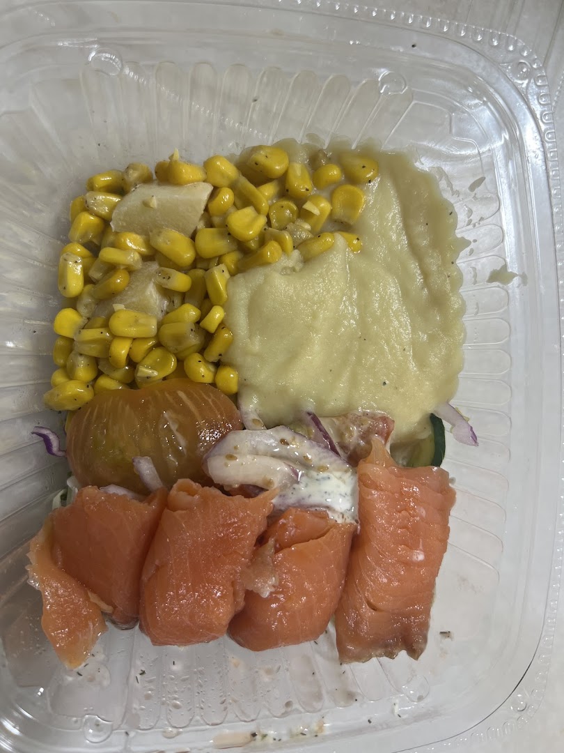 A fresh meal in a clear container featuring smoked salmon, corn, mashed potatoes, and mixed vegetables.