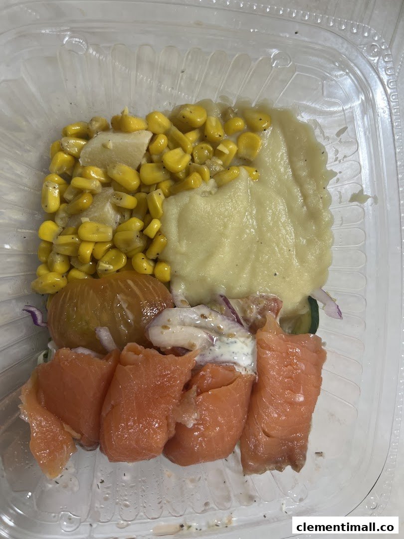 A fresh meal in a clear container featuring smoked salmon, corn, mashed potatoes, and mixed vegetables.