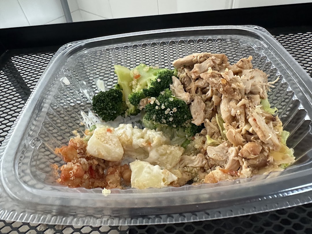 A healthy, customizable meal in a clear plastic container, featuring chicken, broccoli, quinoa, and other fresh ingredients.