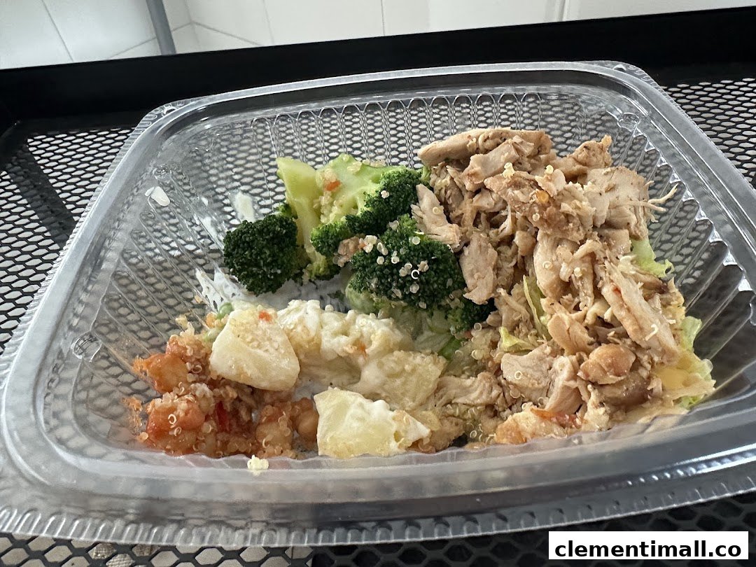 A healthy, customizable meal in a clear plastic container, featuring chicken, broccoli, quinoa, and other fresh ingredients.
