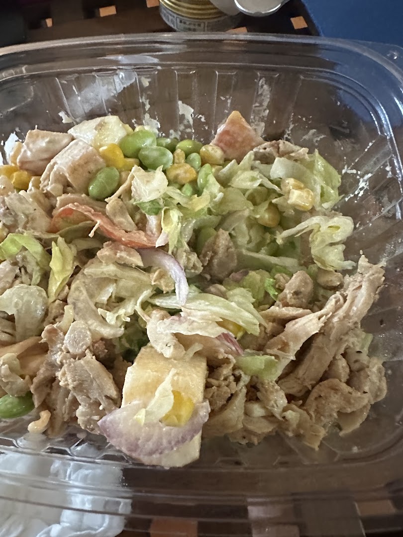 A close-up of a fresh salad bowl with chicken, lettuce, corn, edamame, and red onion in a clear plastic container.