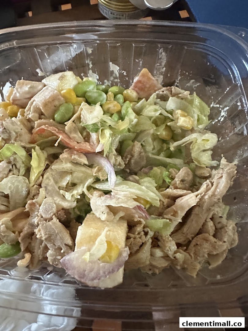 A close-up of a fresh salad bowl with chicken, lettuce, corn, edamame, and red onion in a clear plastic container.