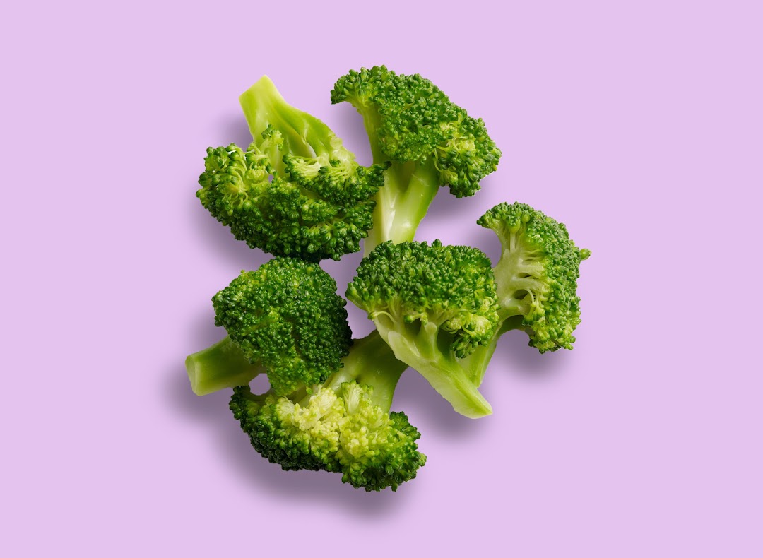 Fresh green broccoli florets are scattered on a light purple background.