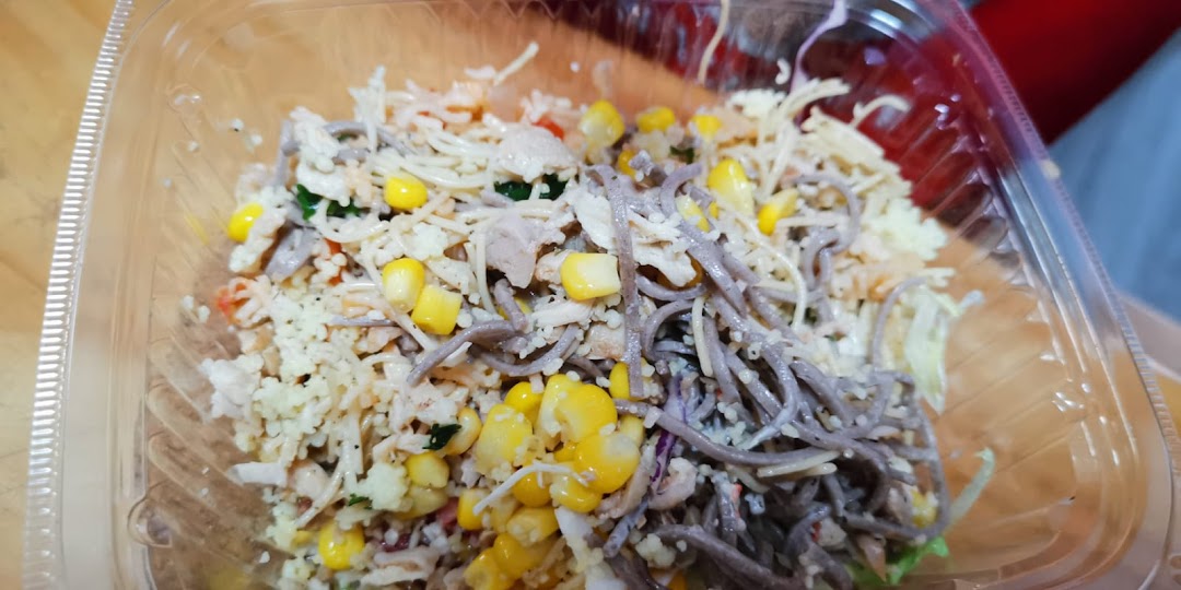 A close-up of a vibrant, customizable food bowl featuring noodles, corn, and protein in a clear container.