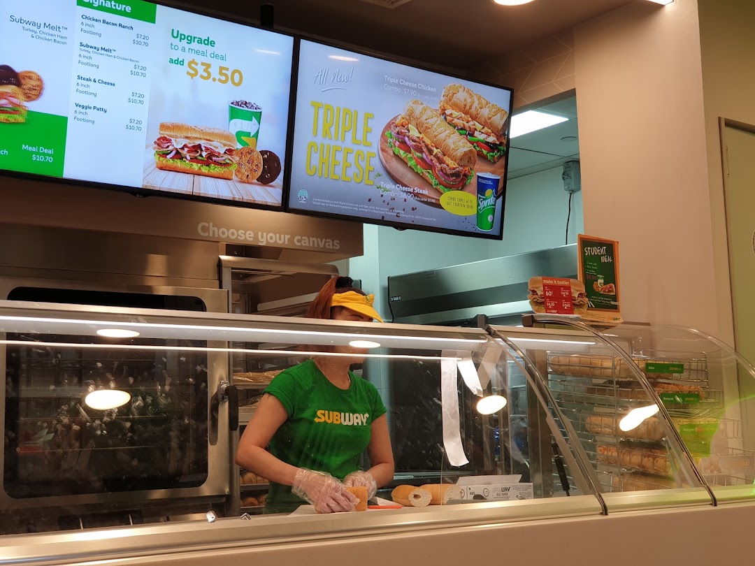 A Subway employee prepares a sandwich behind the counter with digital menu boards displaying meal options.