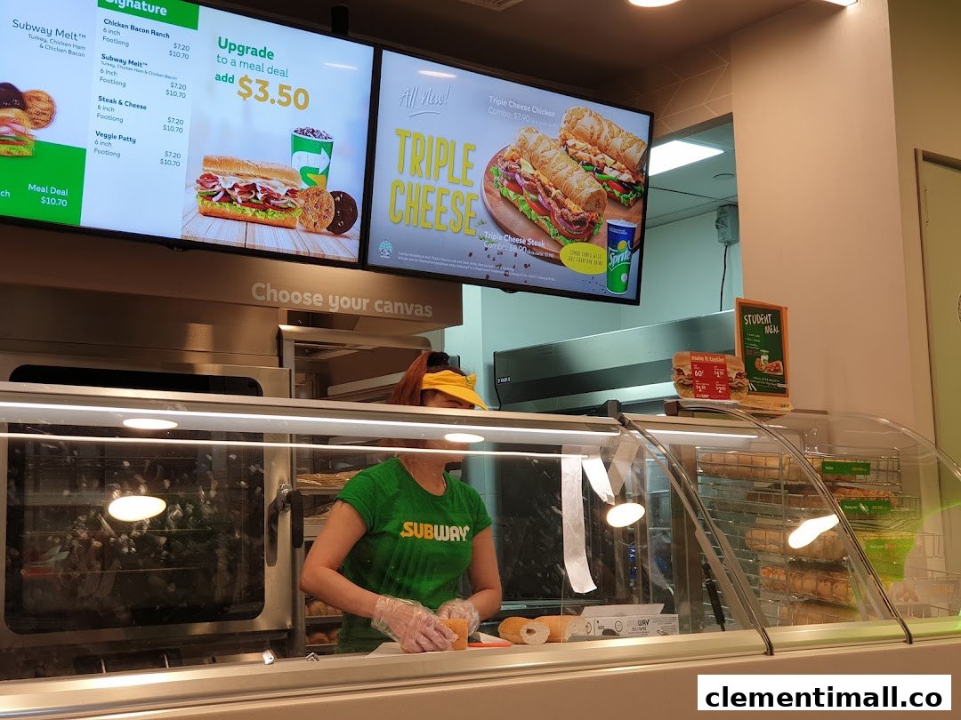 A Subway employee prepares a sandwich behind the counter with digital menu boards displaying meal options.