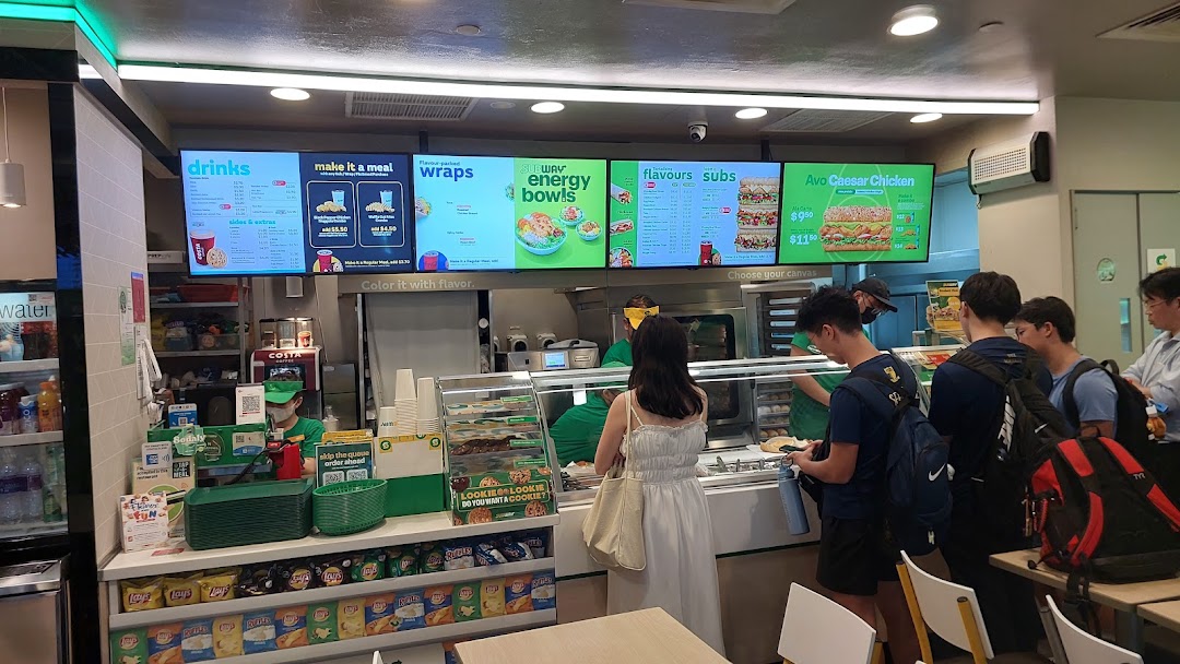 Customers ordering at the counter of a Subway restaurant with digital menu boards displaying various offerings.