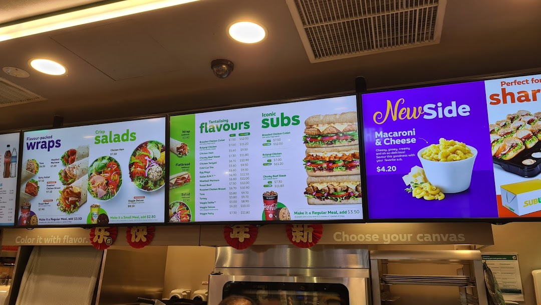 A digital menu board at Subway displaying various subs, wraps, salads, and sides with prices.