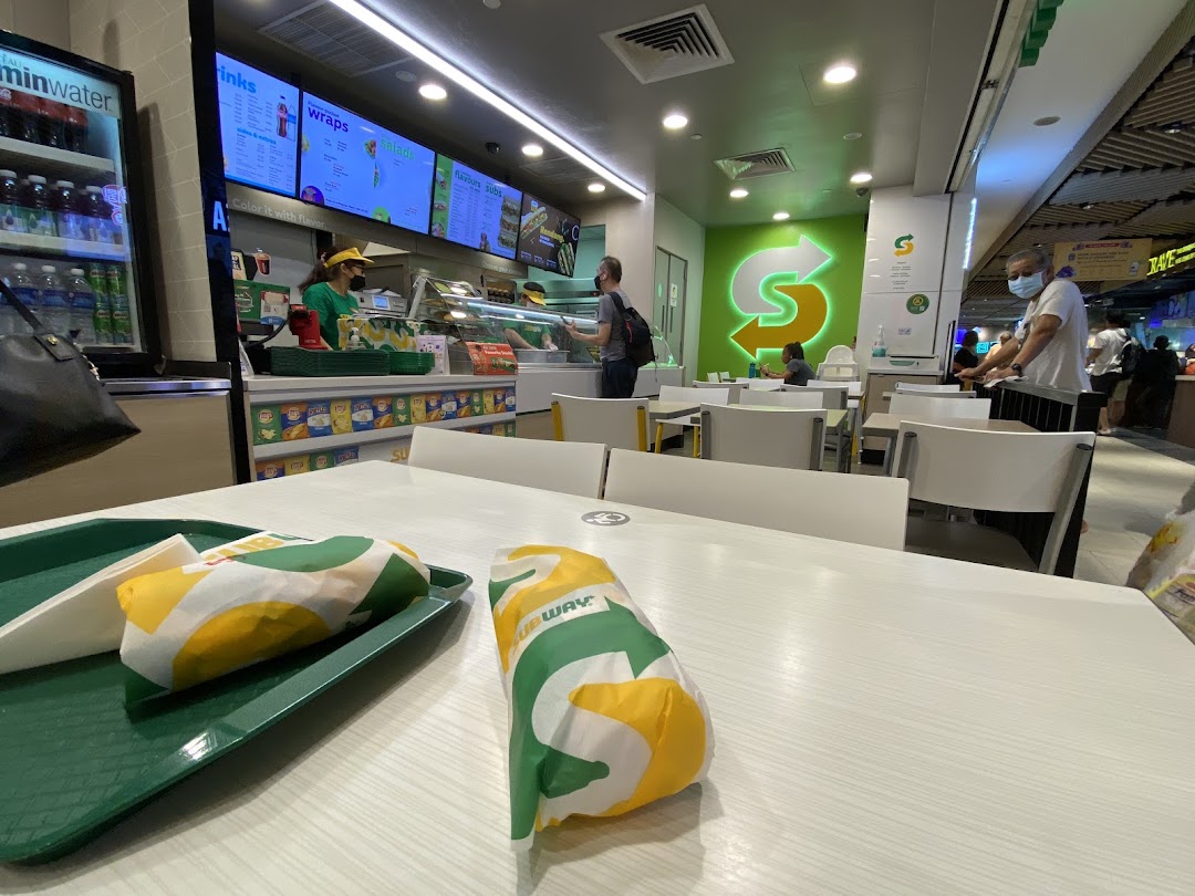 Interior view of a Subway restaurant with customers, staff, and two wrapped subs on a table.
