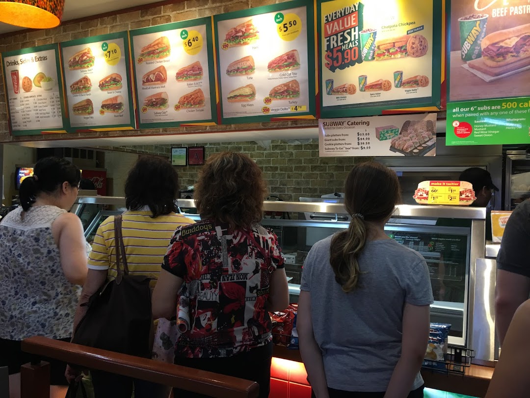 Customers queueing at the counter of a Subway restaurant, with large menu boards displaying various subs and deals.