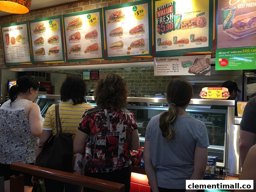 Customers queueing at the counter of a Subway restaurant, with large menu boards displaying various subs and deals.