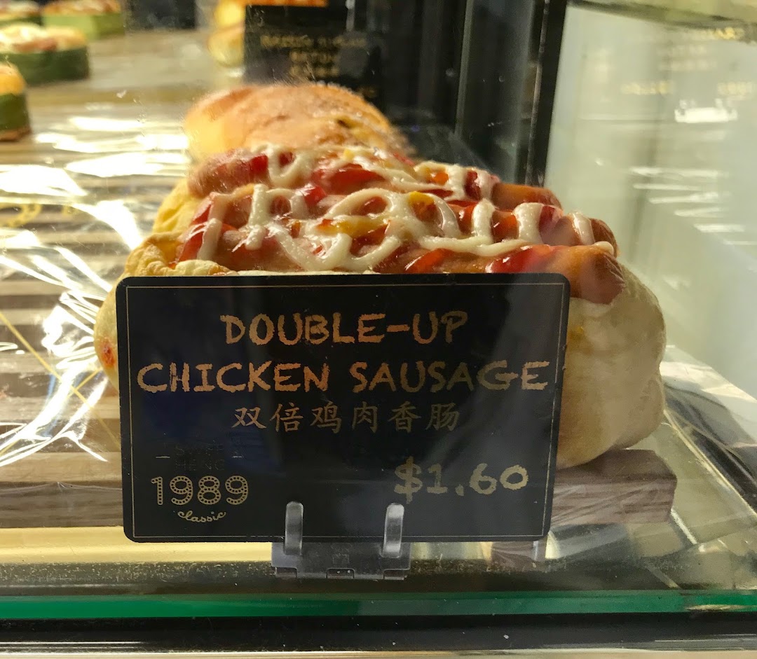 A close-up of a 'Double-Up Chicken Sausage' bun with a price tag in a display case.