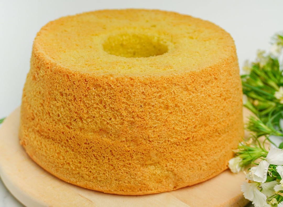 A golden-brown chiffon cake on a wooden board with white flowers.
