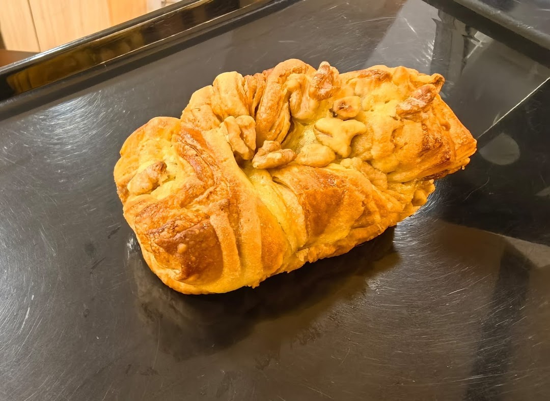 A golden-brown pastry, possibly a bread or bun, topped with walnuts, on a dark baking tray.